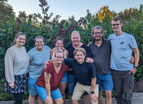 Auf dem Foto sind die acht Teammitglieder zu sehen. Im Hintergrund sind Büsche. Alle lächeln fröhlich in die Kamera.