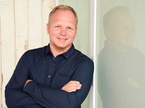 Tim Krenke Auf dem Foto ist Tim Krenke. Er hat blond-rötliches Haar, einen kurzen Bart und lächelt. Er trägt ein dunkelblaues Hemd und lehnt an einer hellen Hauswand.